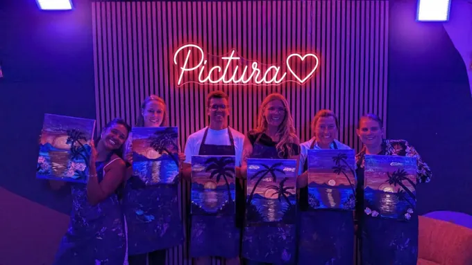A group of adults holding up their finished canvases under the pink Pictura neon sign
