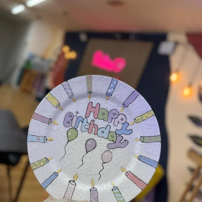 A plate painted with ‘Happy Birthday’, candles and balloons, held up in the studio